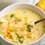 A bowl of chicken broccoli cheddar soup with a spoon resting inside, revealing visible chunks of chicken, broccoli, and melted cheddar.