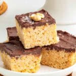 A stack of four chocolate peanut butter Rice Krispie treats on a plate, each with a glossy chocolate top and lightly salted chopped peanuts for garnish.