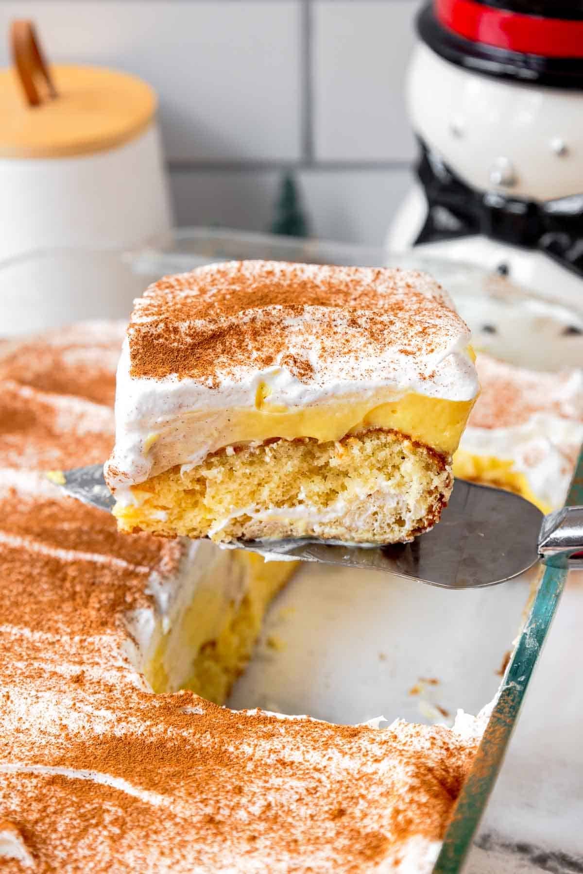 A slice of eggnog poke cake being lifted from a glass baking dish, showing layers of butter cake, eggnog pudding, and whipped topping dusted with cinnamon and nutmeg.