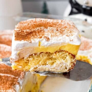 Slice of eggnog poke cake being lifted from a baking dish showing butter cake layer, eggnog pudding layer, and whipped topping layer.