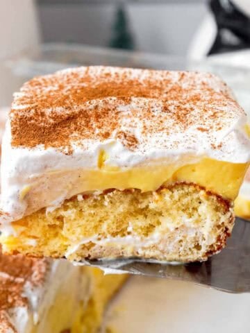 Slice of eggnog poke cake being lifted from a baking dish showing butter cake layer, eggnog pudding layer, and whipped topping layer.