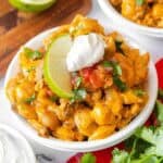 Small white bowl filled with taco mac and cheese, garnished with sour cream, salsa, chopped cilantro, and a lime slice.