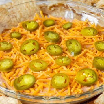 A glass pie dish filled with cream cheese and salsa dip, topped with shredded cheddar cheese and sliced jalapeños, surrounded by tortilla chips.