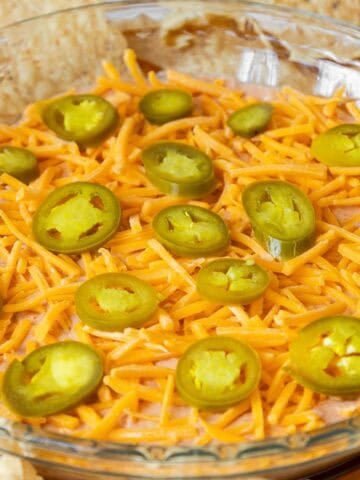 A glass pie dish filled with cream cheese and salsa dip, topped with shredded cheddar cheese and sliced jalapeños, surrounded by tortilla chips.