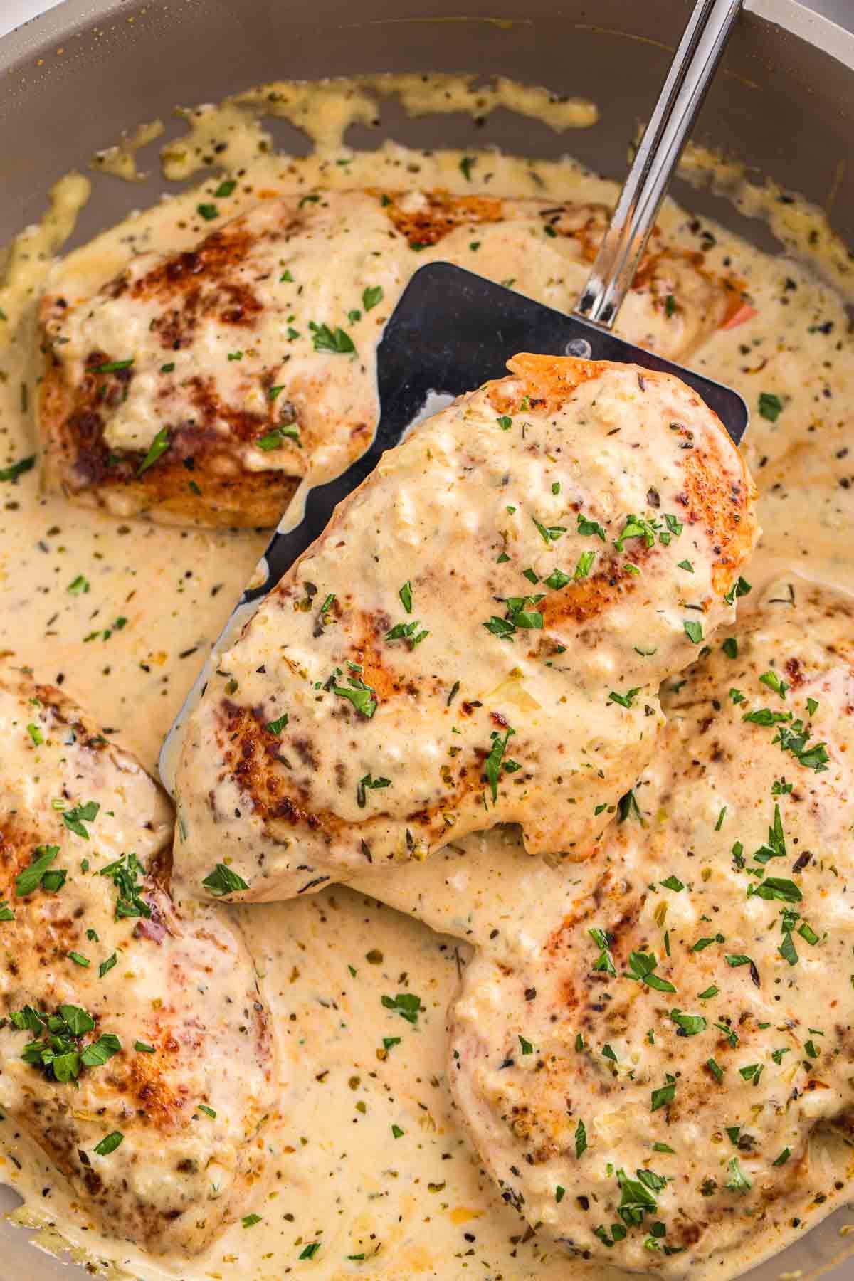Chicken breasts in a skillet covered in creamy garlic sauce and topped with chopped parsley. A spatula lifts one piece to show the sauce coating the chicken.
