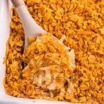 Serving spoon scooping baked hash brown casserole from a baking dish, showing the creamy interior, shredded potatoes, and crisp golden corn flake topping.