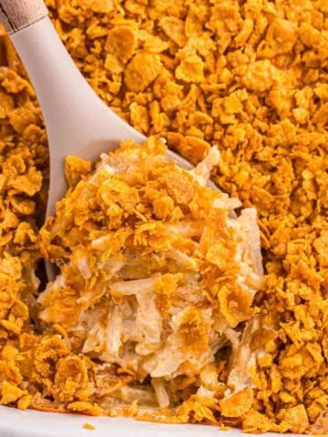 Serving spoon scooping baked hash brown casserole from a baking dish, showing the creamy interior, shredded potatoes, and crisp golden corn flake topping.
