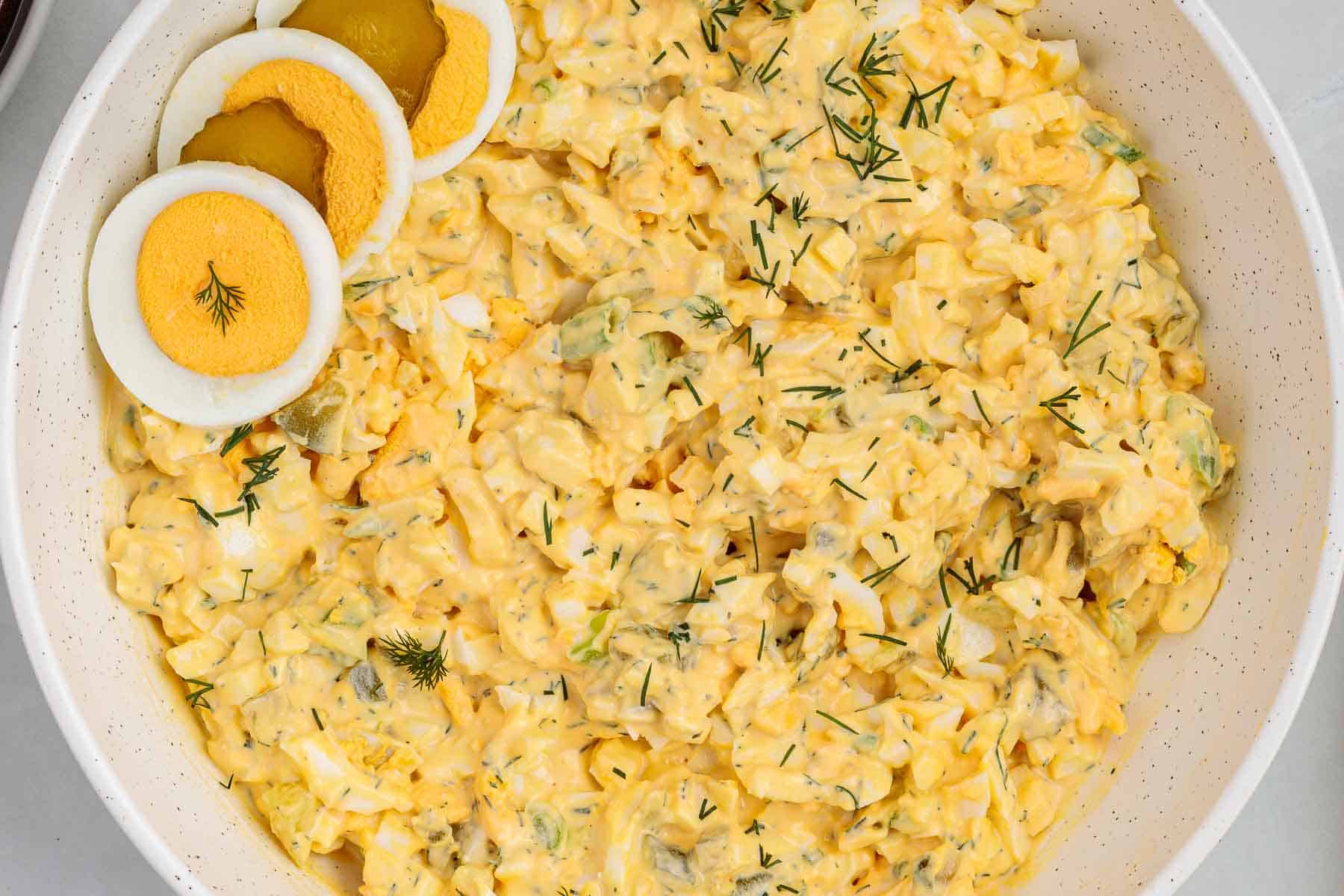 Overhead view of a large bowl filled with dill pickle egg salad mixed with chopped eggs, pickles, and fresh dill, with sliced hard boiled eggs arranged on top.