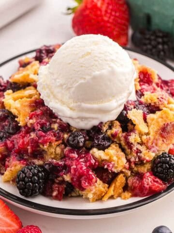 Berry cobbler with cake mix topped with vanilla ice cream on a plate with fresh berries nearby.