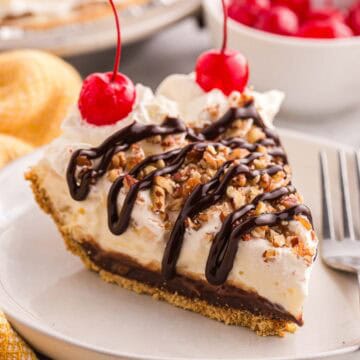 Slice of banana split pie topped with whipped cream, chopped pecans, chocolate syrup, and cherries.