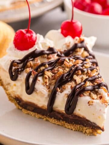 Slice of banana split pie topped with whipped cream, chopped pecans, chocolate syrup, and cherries.