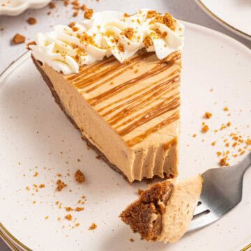 Close view of a slice of no bake Biscoff cookie butter pie with a thick creamy filling and crumb crust. Whipped cream and crushed Biscoff cookies top the slice with cookie butter drizzled across the surface. A bite is taken out and sitting on a fork.