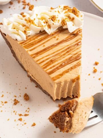 Close view of a slice of no bake Biscoff cookie butter pie with a thick creamy filling and crumb crust. Whipped cream and crushed Biscoff cookies top the slice with cookie butter drizzled across the surface. A bite is taken out and sitting on a fork.