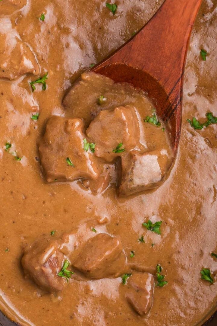 Tender beef tips surrounded by thick brown gravy in the slow cooker. A wooden spoon lifts several pieces of beef to show the texture and consistency of the gravy.