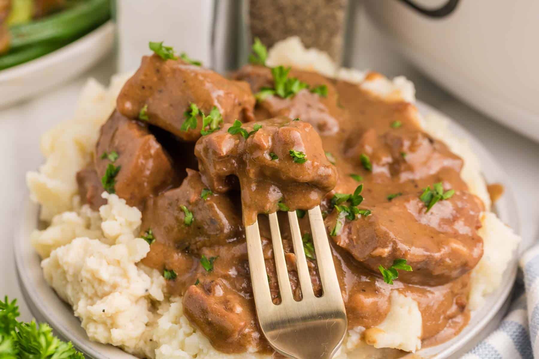 Slow cooker beef tips and gravy served over mashed potatoes. A fork holds one gravy coated piece of tender beef above the serving.