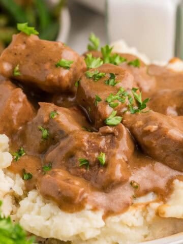 Close view of tender slow cooker beef tips covered in rich brown gravy and served over mashed potatoes. Chopped parsley is sprinkled over the beef before serving.