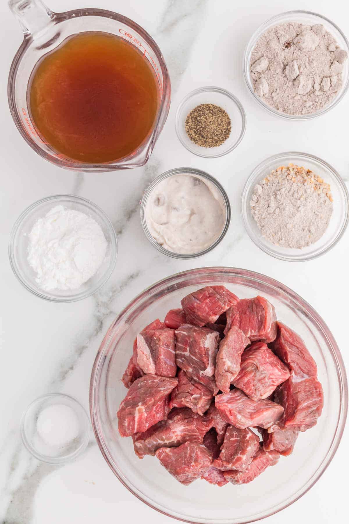 Ingredients for slow cooker beef tips and gravy arranged before cooking. Cubed raw beef is shown with beef broth. Cream of mushroom soup. Onion soup mix. Brown gravy mix. Cornstarch. Salt. And black pepper.