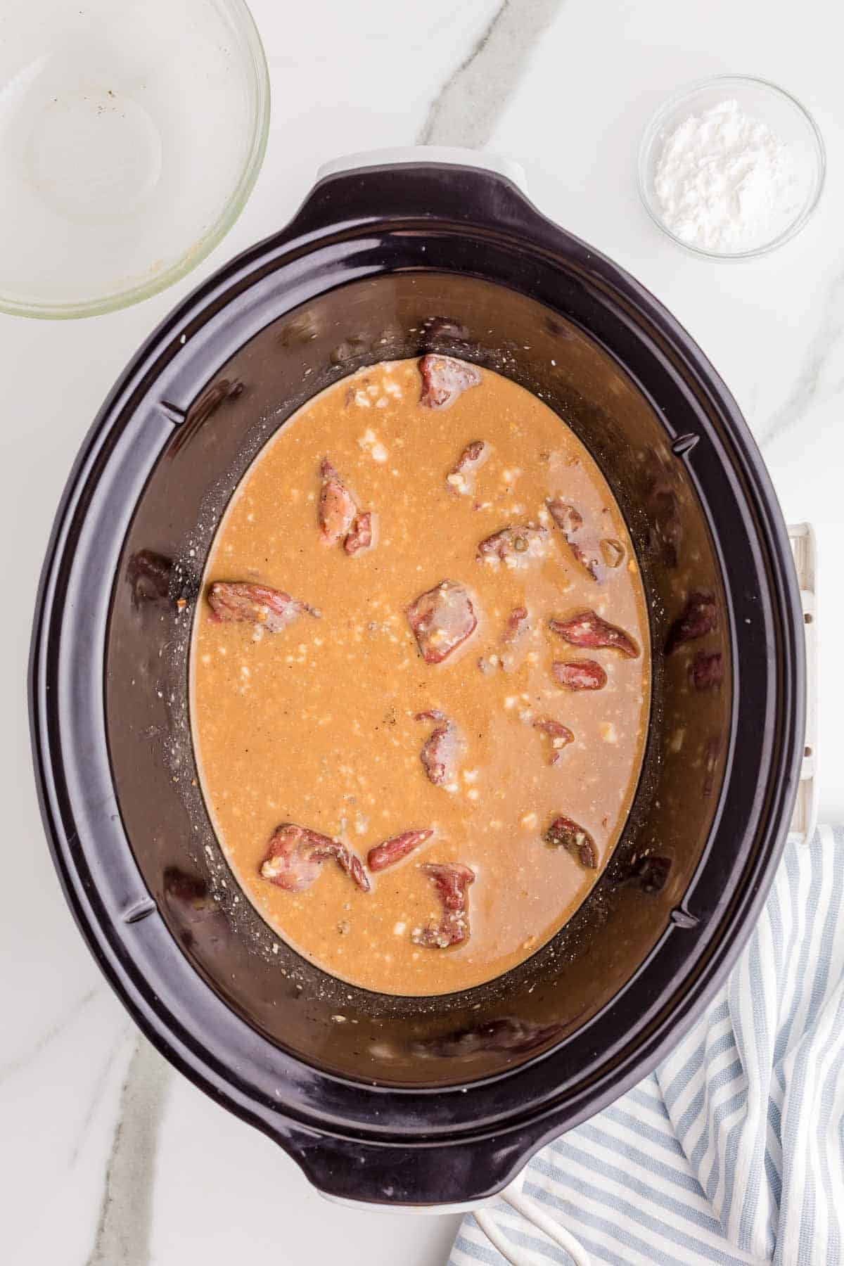 Seasoned beef pieces added to the gravy mixture in the slow cooker. The beef is spread throughout the liquid before the slow cooking begins.