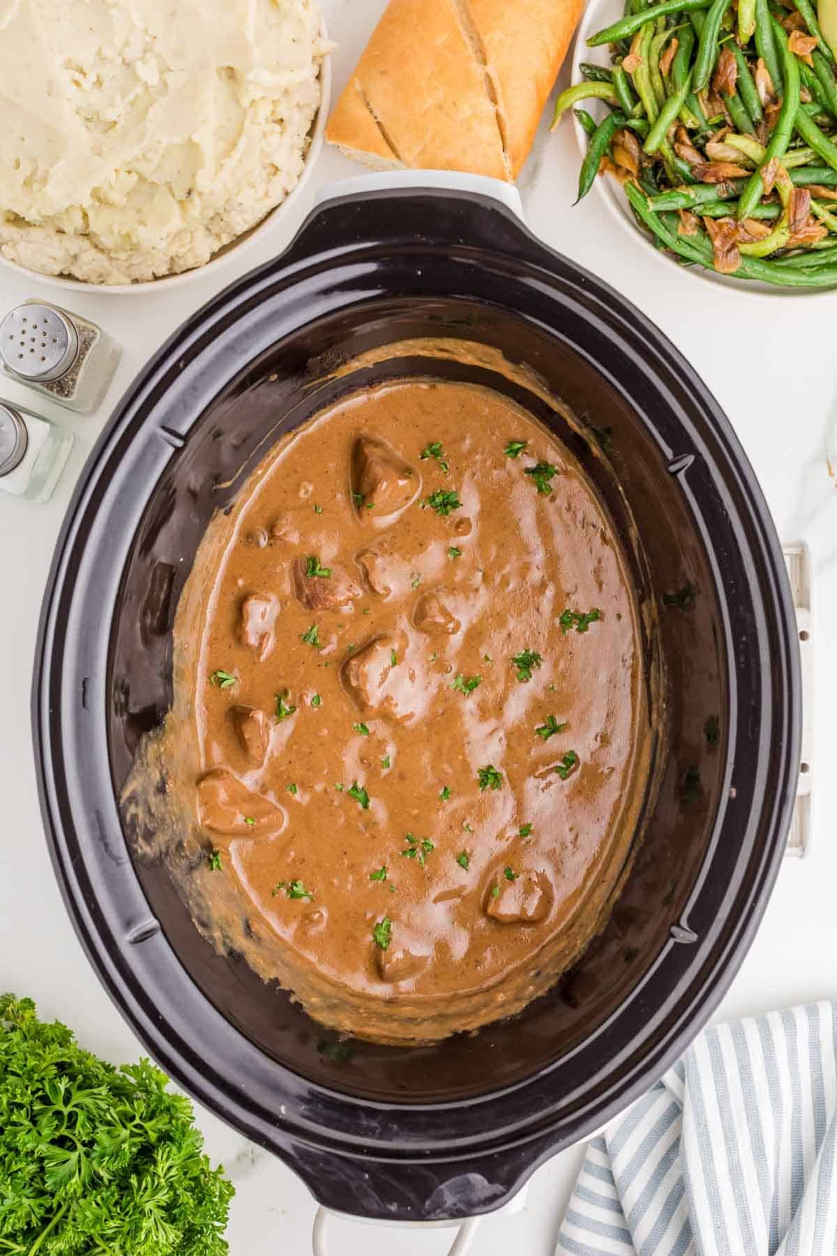Finished slow cooker beef tips in thick brown gravy with chopped parsley scattered over the top. Mashed potatoes and green beans are ready alongside the slow cooker for serving.