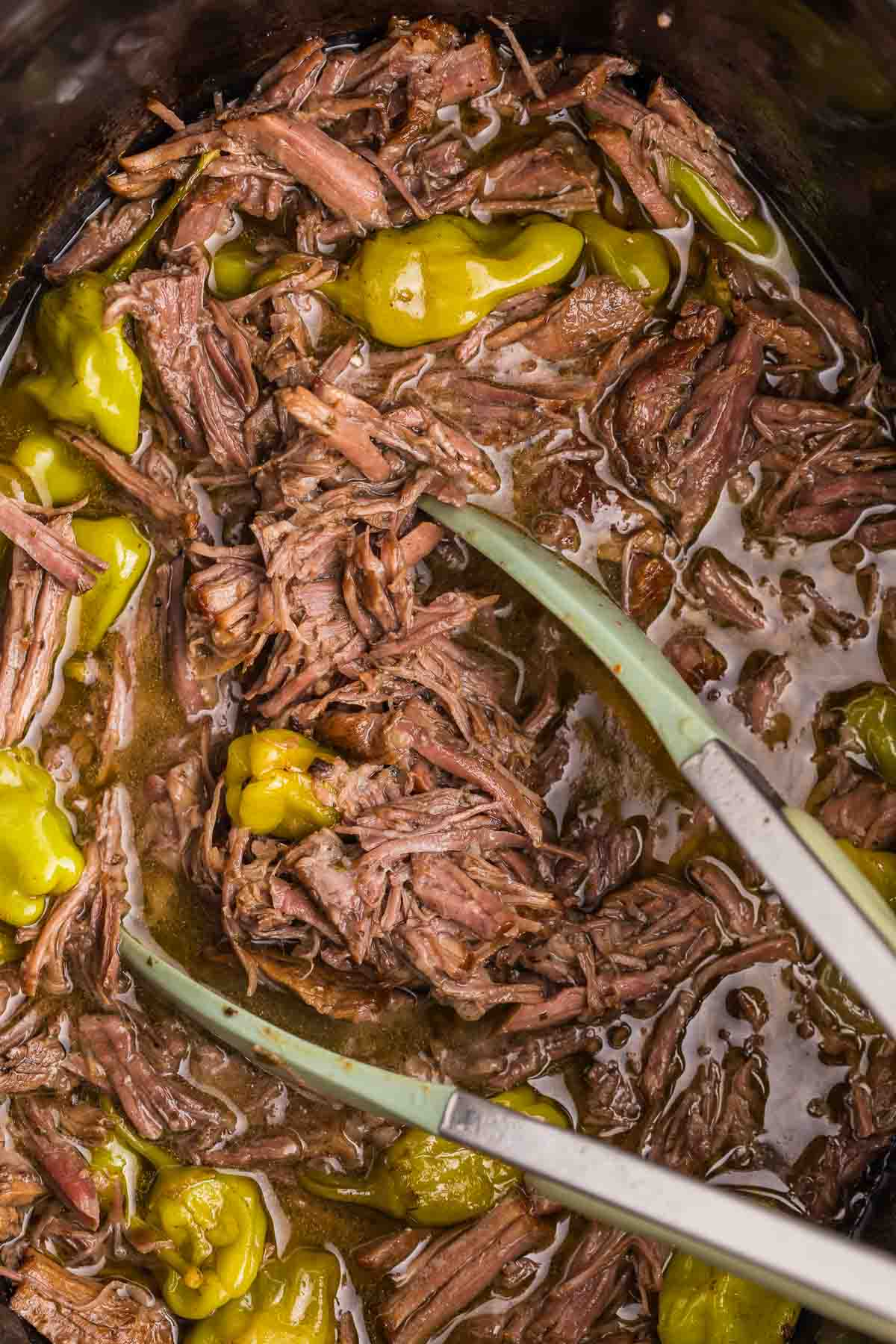 Shredded Mississippi pot roast sits in its rich cooking juices with whole pepperoncini peppers in the slow cooker. Two silicone tipped tongs are positioned in the beef after it has been pulled apart.